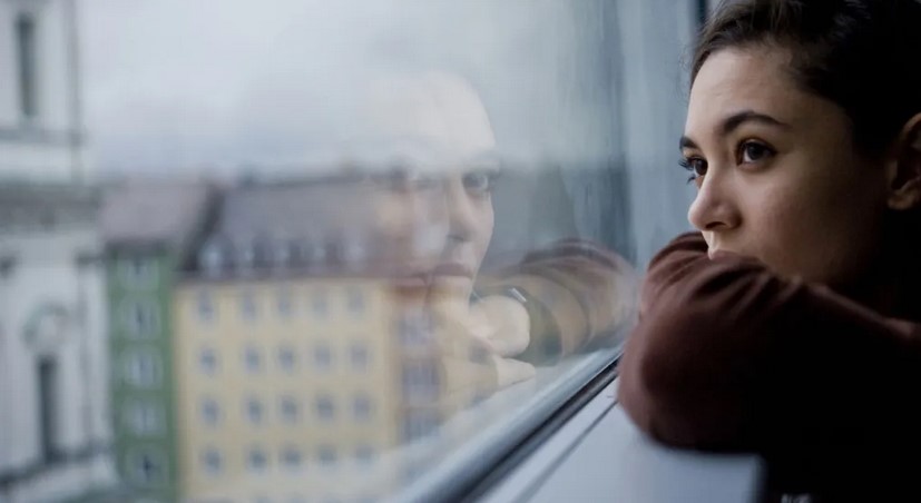 Color photograph of a young woman's profile against a window looking out on a rainy day with cars going by